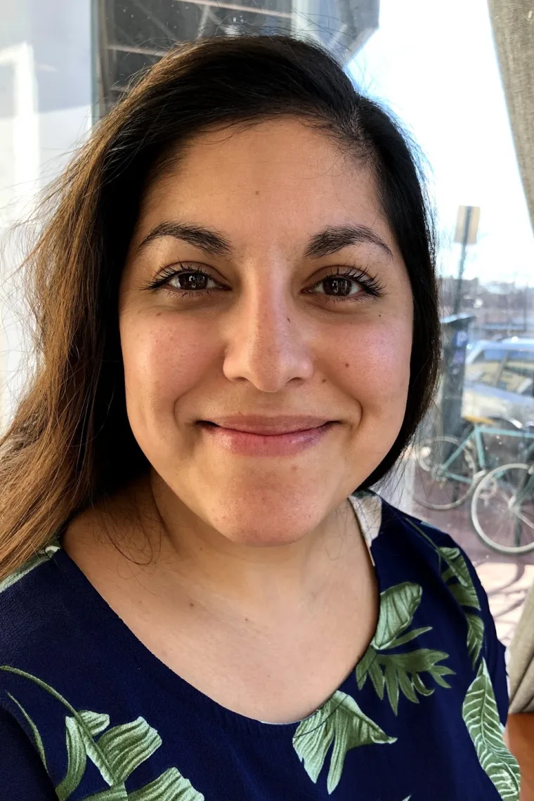 Person smiling in close-up, wearing a blue top with green leaf patterns. A bicycle and building are visible in the background.