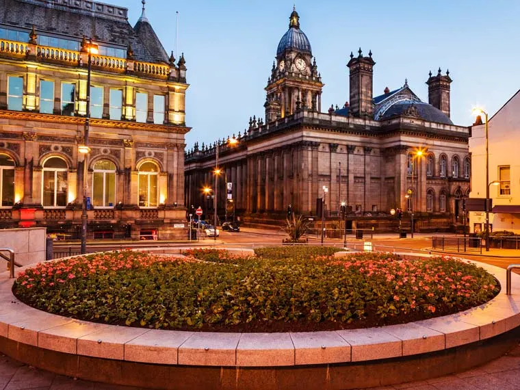 Historic building with clock tower and dome, lit warmly in early evening. Foreground features circular flower bed with surrounding railings.