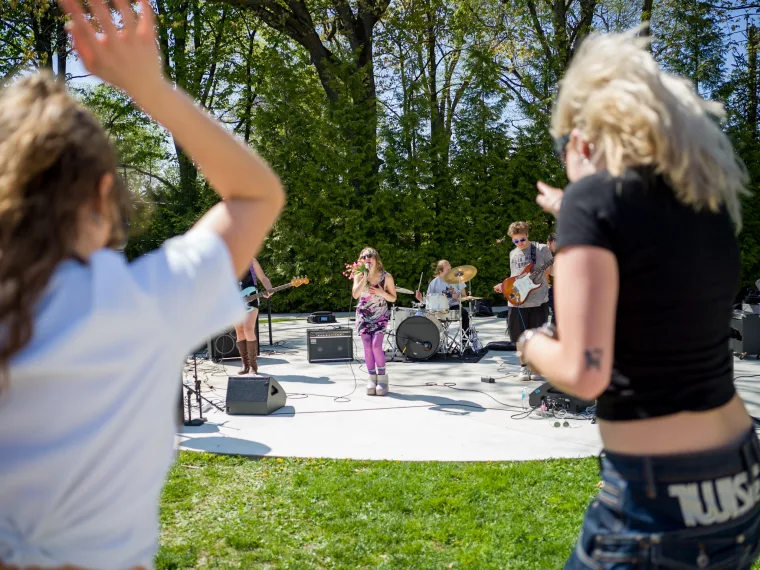 People dance in the foreground while a band performs on an outdoor stage surrounded by trees.