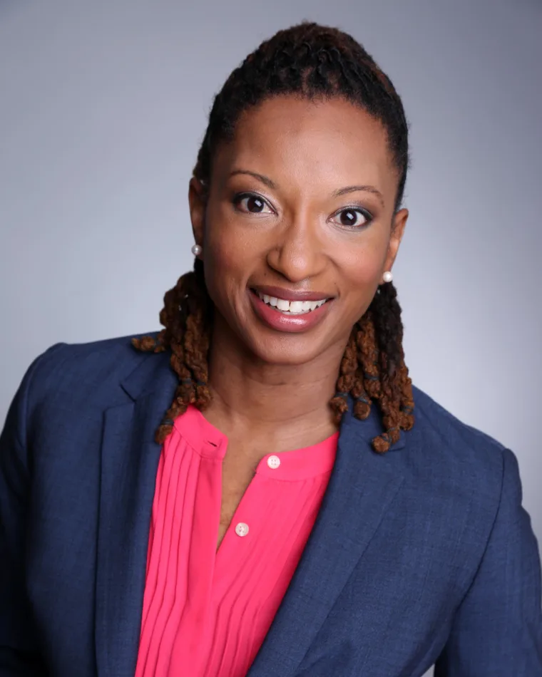 Person with braided hair smiles, wearing a navy blazer and pink shirt, against a gray background.