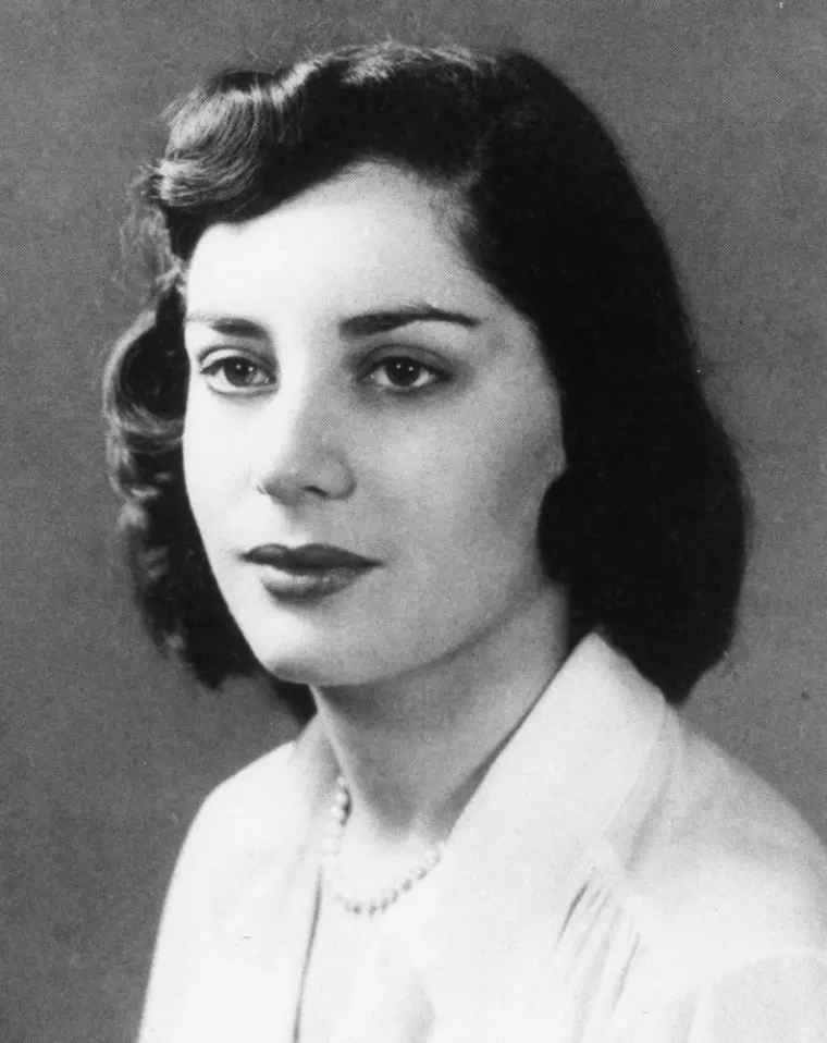Person with short, wavy hair wearing a light-colored blouse and pearl necklace in a vintage black and white portrait.