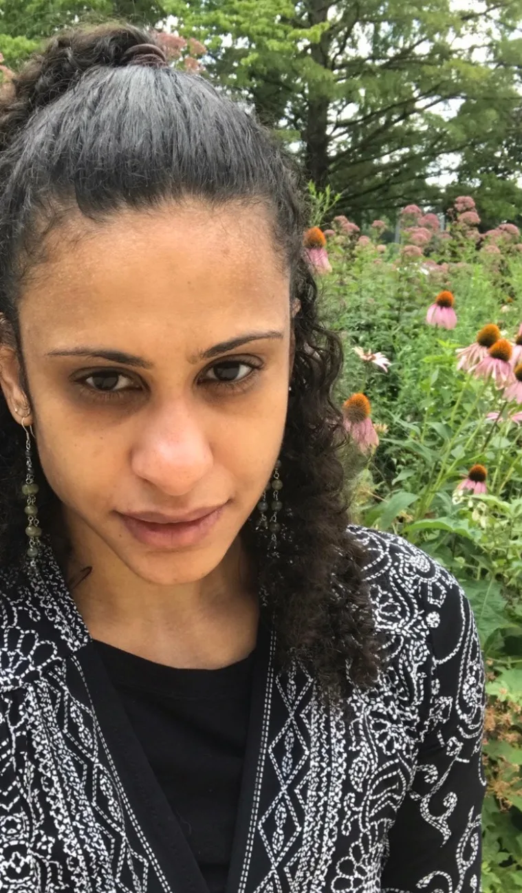 Person with curly hair in patterned top, standing in a garden with purple and orange flowers.