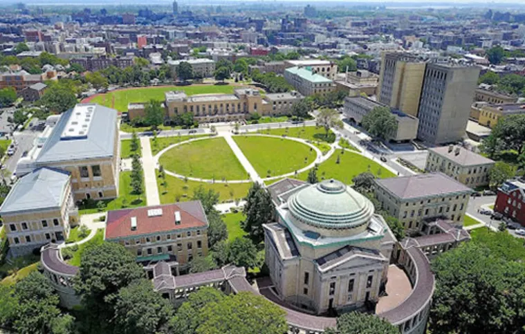 Aerial view of a college campus with a circular lawn, surrounded by academic buildings, pathways, and trees, set against a cityscape backdrop.