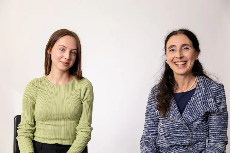Two people sitting and smiling; one wearing a light green sweater and the other in a blue patterned jacket, against a plain background.