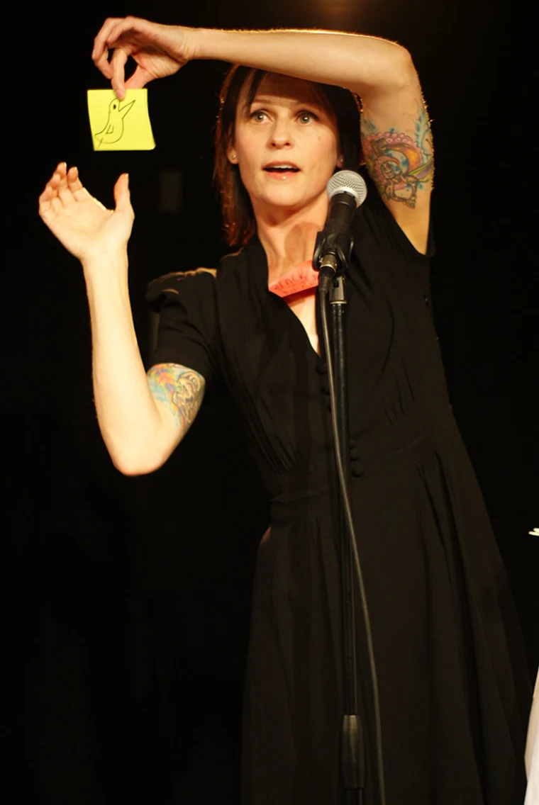 Person with tattoos holding a yellow sticky note with a bird drawing on stage, speaking into a microphone.