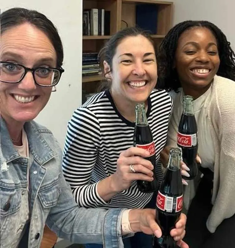 Three people smiling and holding glass Coca-Cola bottles, standing close together in a room with shelves in the background.