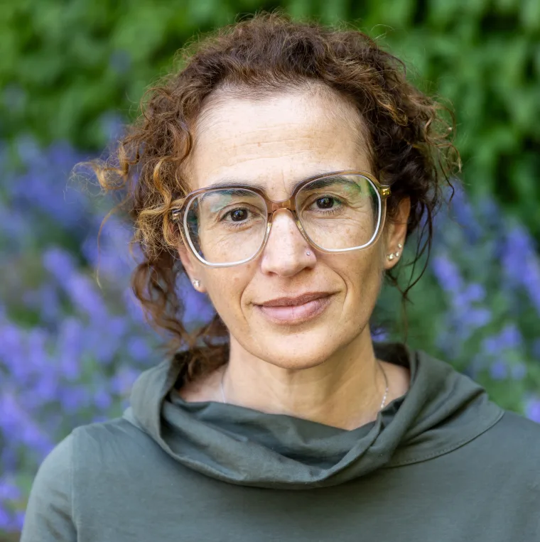 Person with curly hair and glasses smiles slightly, standing in front of a blurred background of green leaves and purple flowers.