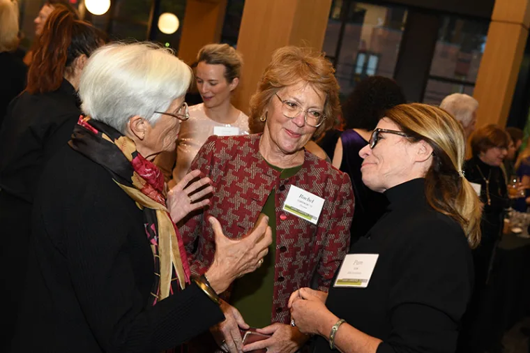 Three people are engaged in conversation at an event, wearing name tags. Others are socializing in the background.