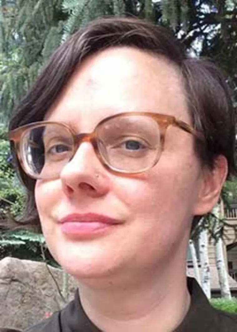 Person with short dark hair and glasses, standing outdoors with trees in the background.