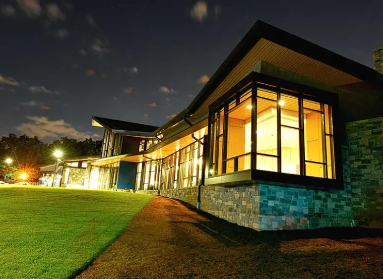 Modern building with large windows illuminated at night, surrounded by a lawn.