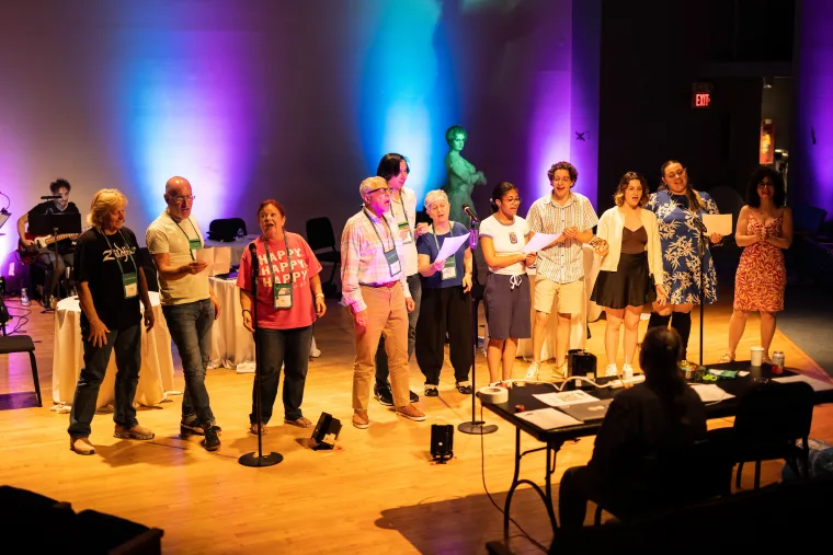 A group of people standing on stage with microphones, performing under colorful stage lighting. A pianist and table with judges are in the foreground.