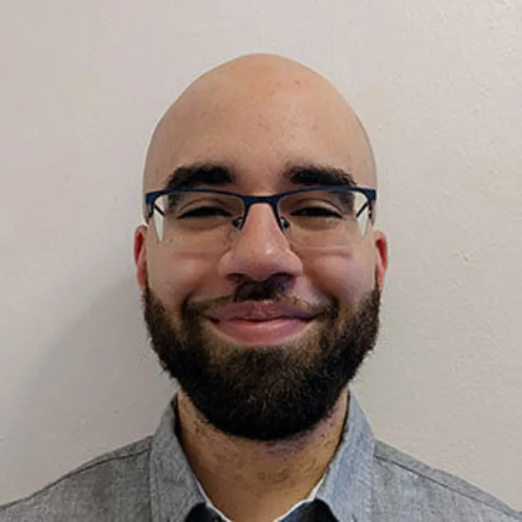Person with glasses and a beard, smiling, wearing a gray collared shirt, standing against a plain white background.
