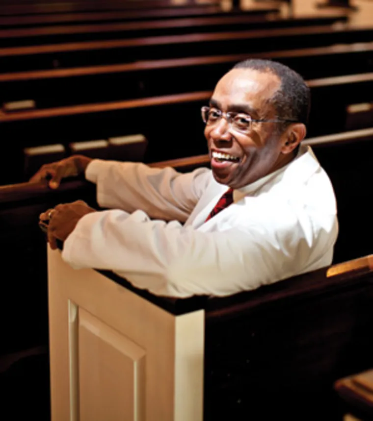 Person wearing glasses and a white jacket, smiling while sitting in a church pew, with rows of benches in the background.