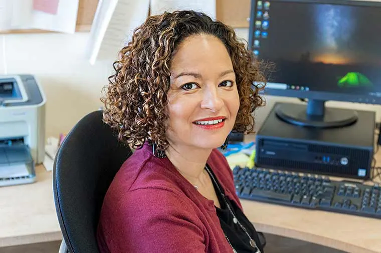 Person with curly hair sitting at a desk, smiling at the camera. A computer monitor and various office supplies are visible in the background.