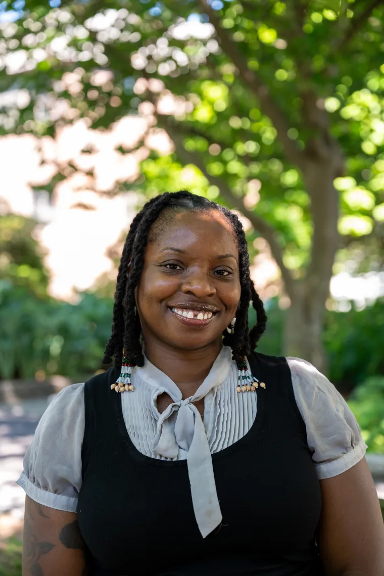 Person smiling outdoors, with braided hair and a black and white outfit, standing in front of blurred greenery and trees.