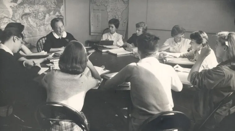 A group of people sitting around a table in a classroom, with maps on the wall, engaging in discussion or study.