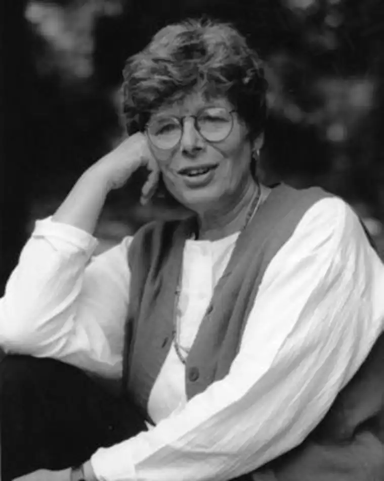A person with short hair and glasses, wearing a white shirt and vest, rests a head on a hand, smiling outdoors.