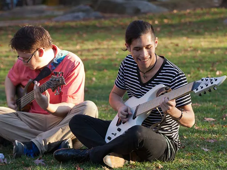 Two people sitting on grass, playing guitars and smiling. One wears a striped shirt, the other a red shirt. Sunlight filters through trees.