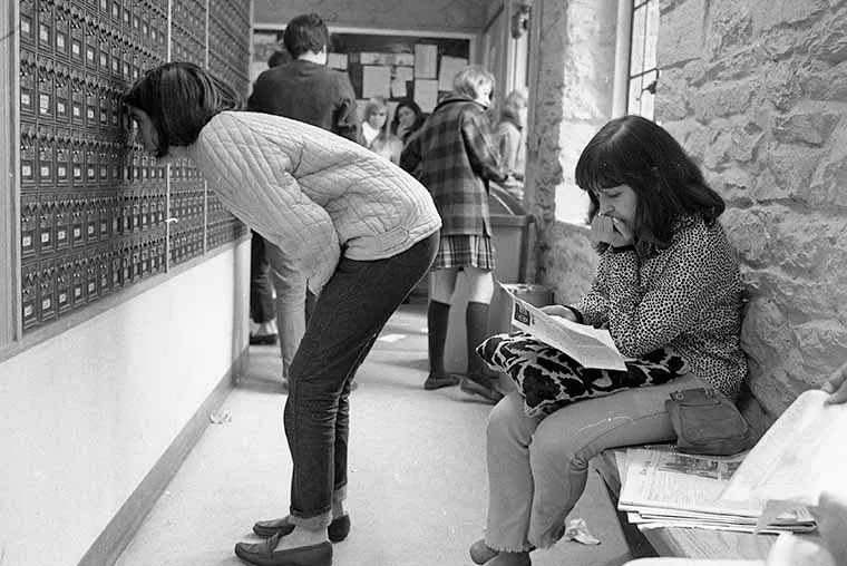 Person looking into a wall of mailboxes, another sitting on a bench reading a letter. Group gathered in the background.