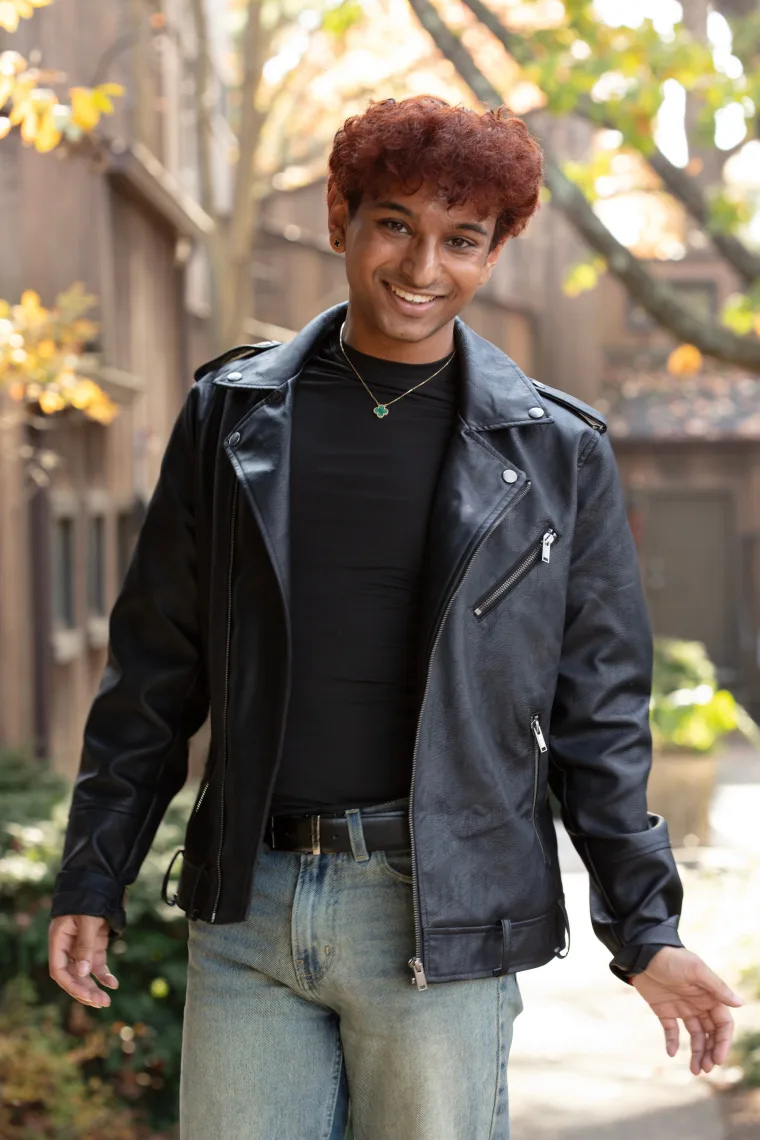 Smiling person with curly hair wearing a black leather jacket and jeans, standing in a sunlit outdoor area with autumn leaves.