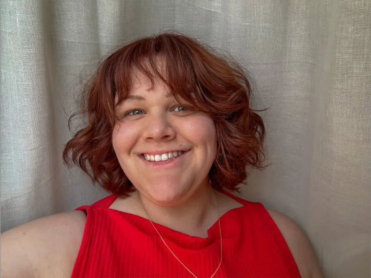 Person with wavy red hair smiling, wearing a red top, against a light-colored curtain background.