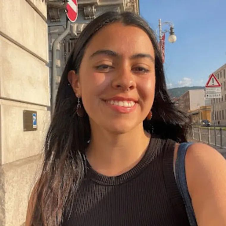 Person smiling outdoors, wearing a black top and earrings, with a sunlit city street and buildings in the background.