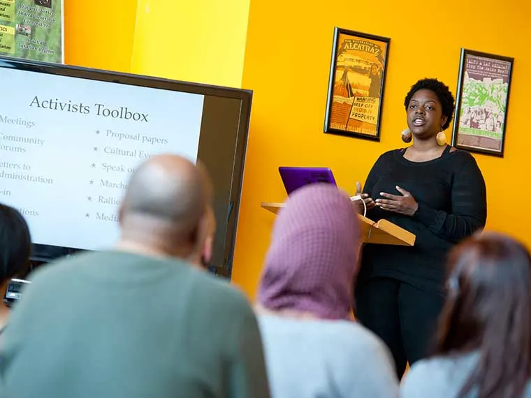 Person presenting on activism with a slide titled "Activists Toolbox" to a small audience in a room with a bright yellow wall.
