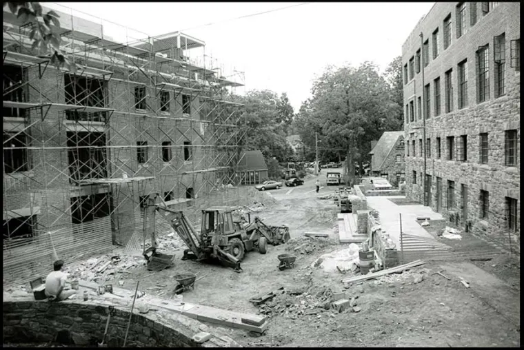 Construction site with scaffolding on a building's facade, a backhoe, and scattered materials. Trees and more buildings in the background.