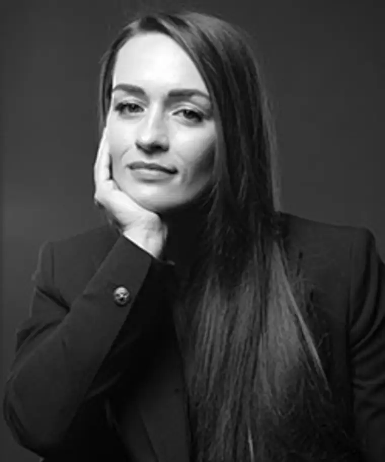 Person with long hair and a relaxed expression, resting their chin on a hand. Wearing a dark suit jacket against a plain background.
