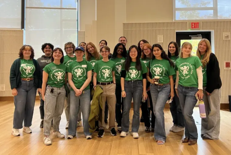 A group of people wearing matching green shirts with a graphic design, standing in a well-lit room with wooden floors and large windows.
