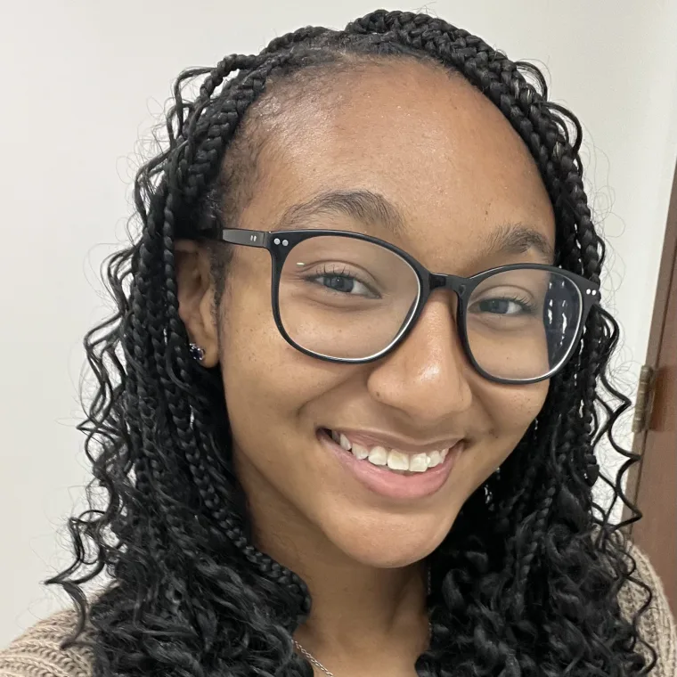 Person smiling, wearing black-rimmed glasses and braided hair, in a well-lit indoor setting.