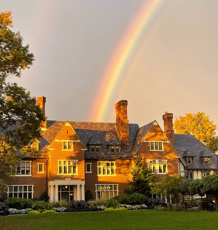 A large brick house with a slate roof is illuminated by warm sunlight. A vibrant rainbow arcs across the sky above, set against a backdrop of trees.