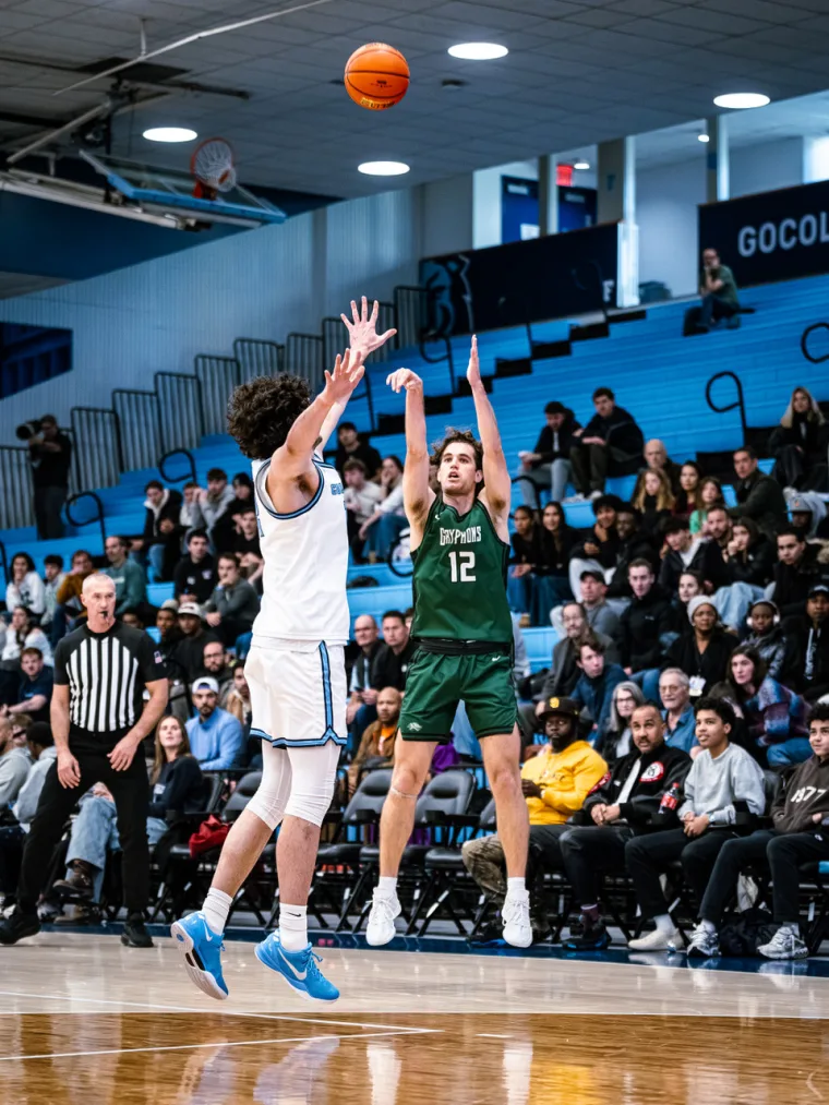 Basketball player in green 12 jumps to block a shot from an opponent in white during a game, with a crowd and referee watching.
