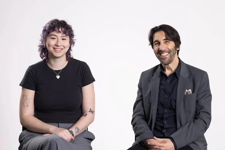 Two people sitting and smiling against a white background. One has colorful hair and tattoos; the other wears a suit with an animal pin.