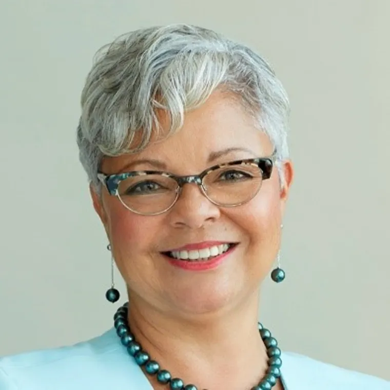 Person with short gray hair, wearing glasses and a teal necklace with matching earrings, smiles against a neutral background.