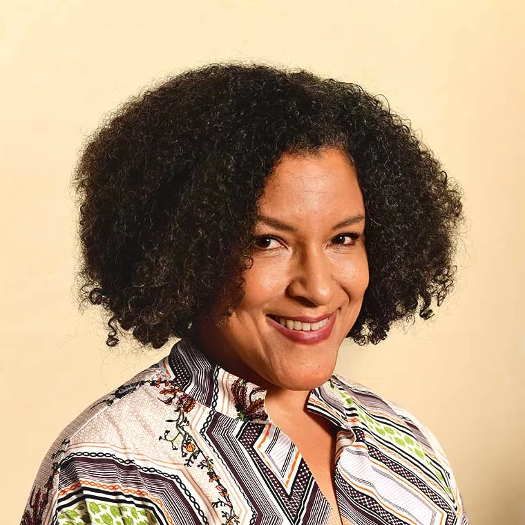 Person with curly hair smiling, wearing a patterned shirt, against a beige background.