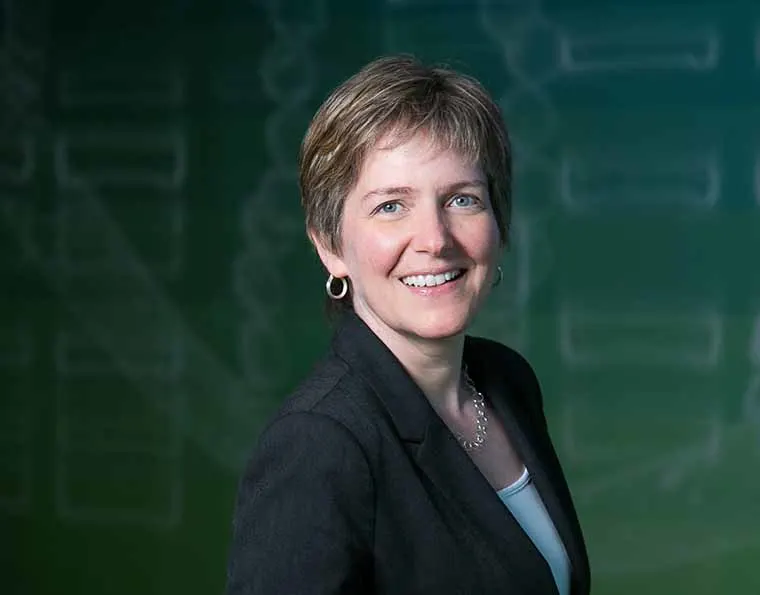 Person smiling while wearing a dark suit, standing against a green background with faint diagram-like patterns.
