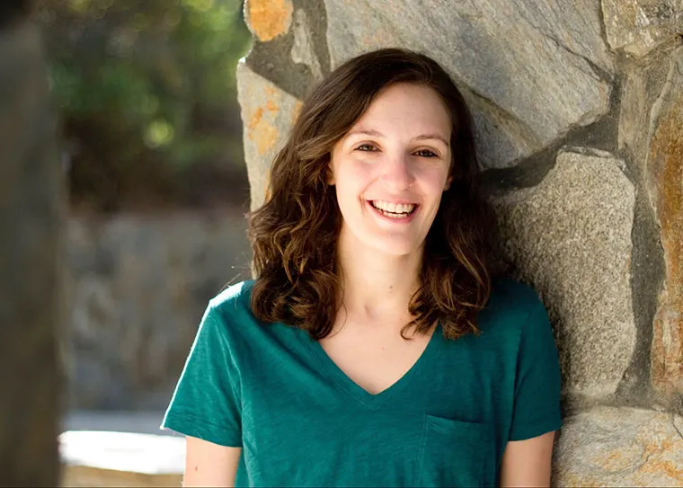 Person with shoulder-length hair smiling, wearing a teal shirt, leaning against a stone wall, with a blurred outdoor background.