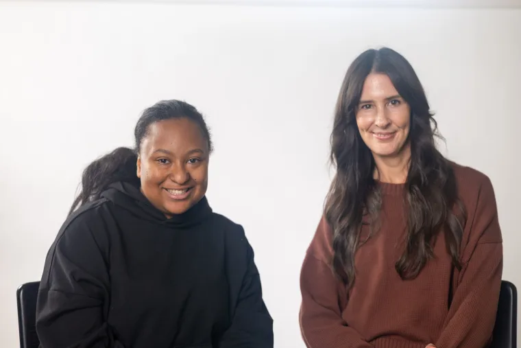 Two people smiling, one wearing a black sweater and the other a brown sweater, seated against a plain white background.