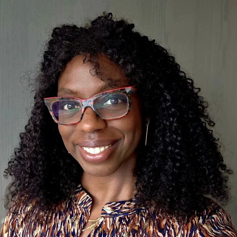 Person with curly hair, wearing glasses and a patterned top, smiles in front of a neutral background.
