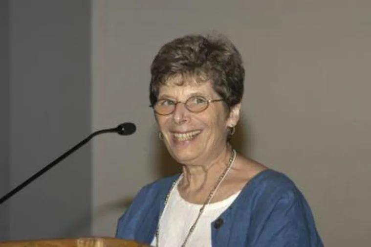A person with short hair and glasses smiles while speaking at a microphone, wearing a blue jacket and white top.