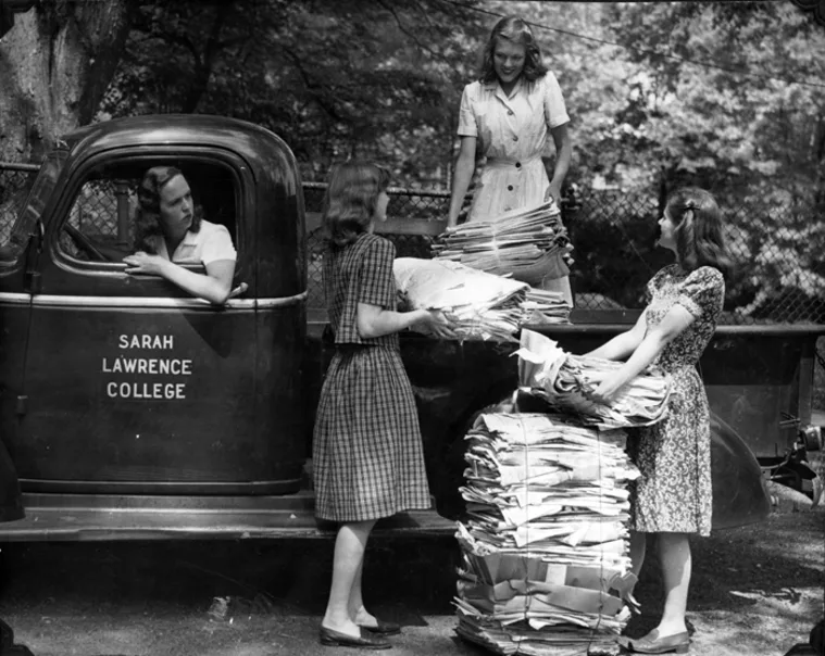 Truck with "Sarah Lawrence College" logo. People loading and stacking large bundles of paper in a wooded area.