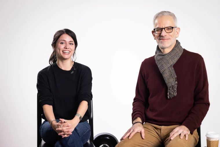 Two people sitting and smiling in a studio. One wears a black sweater and jeans, the other a maroon sweater with a scarf. A coffee cup is nearby.