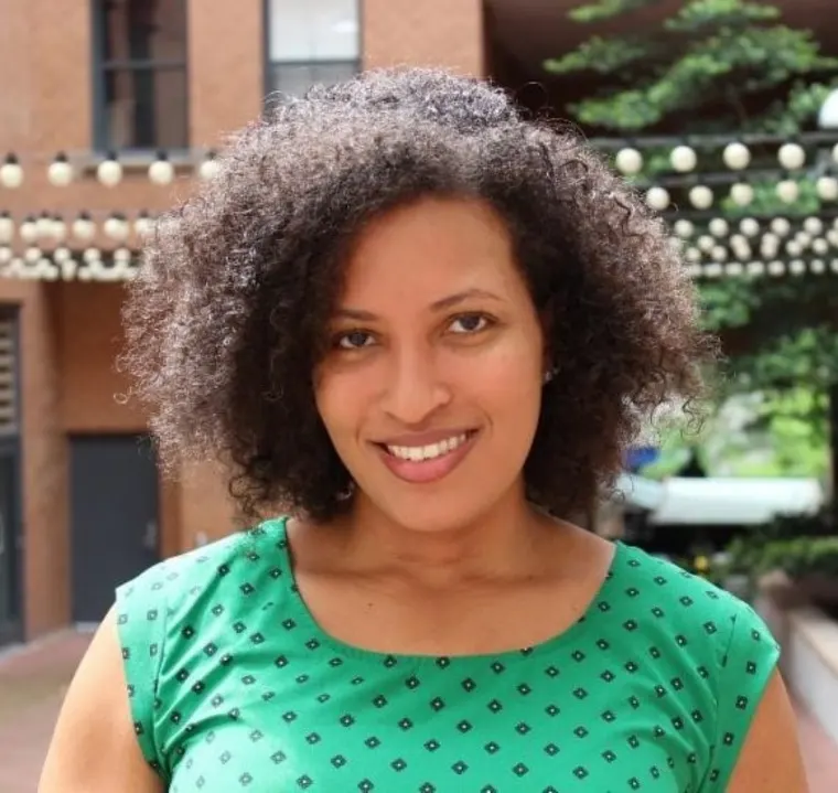 Person smiling outdoors, wearing a green top with a pattern. Background includes brick wall, string lights, and greenery.