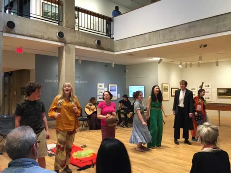 A group of people performs enthusiastically in an art gallery, with musicians playing in the background. Audience members are seated nearby.