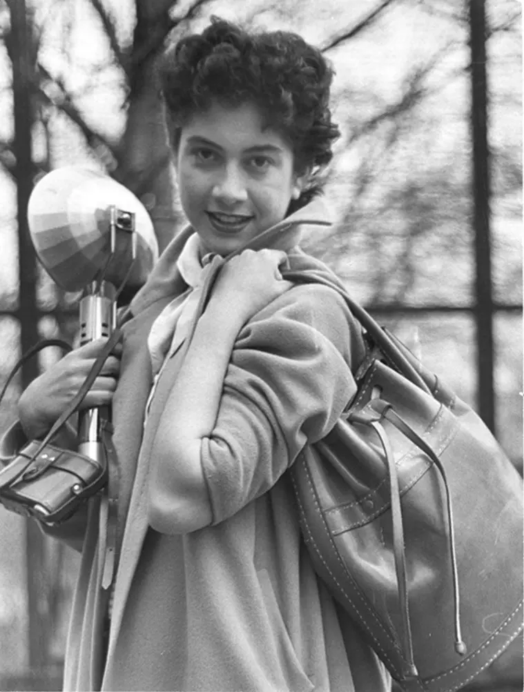 Person smiling, carrying a coat, a bag, and an umbrella with a striped pattern. Trees in the background suggest an outdoor setting.
