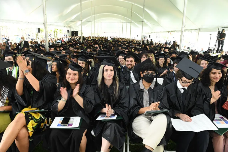 Graduates in caps and gowns sit under a large tent, applauding. Some hold diplomas, and the crowd spans the entire covered area.