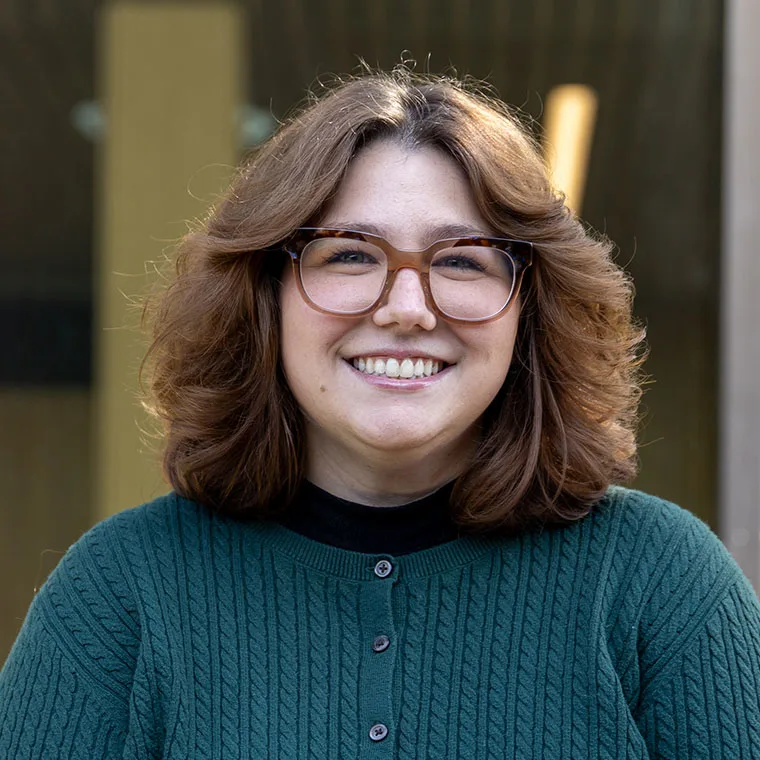 Person with curly hair and glasses smiling, wearing a green sweater, standing outdoors.