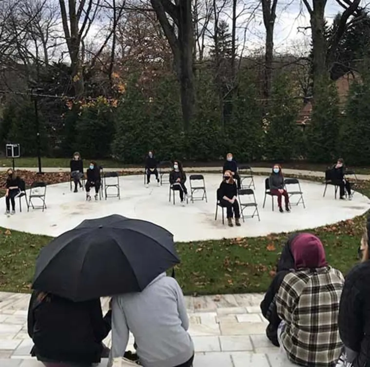 People sitting on chairs spaced apart in an outdoor circular area with masks on. Observers with umbrellas watch from a distance.