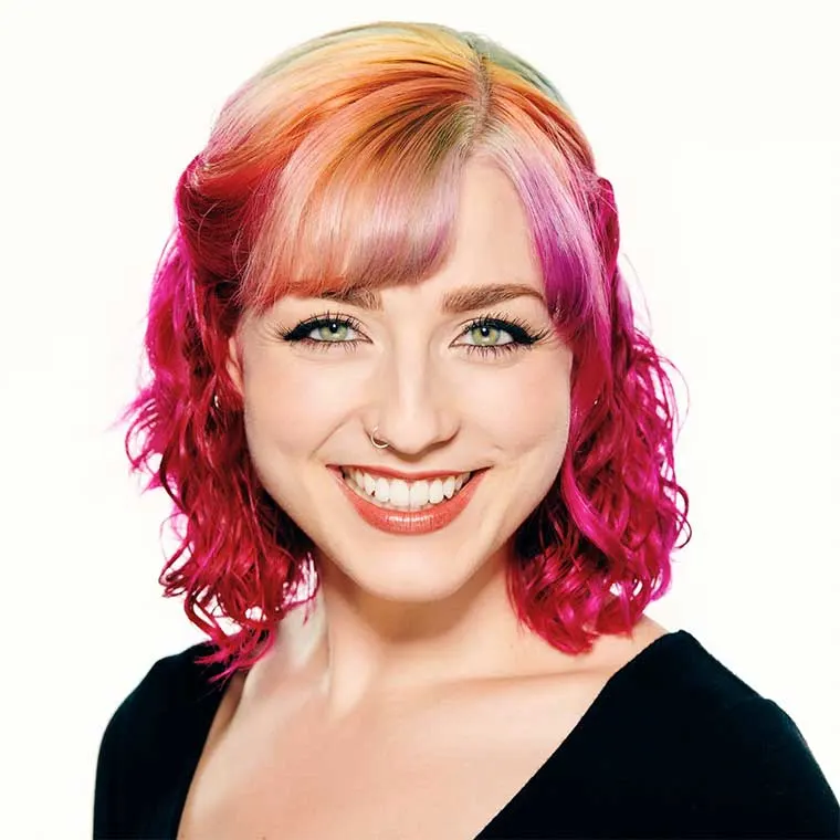 Person with colorful hair, blending pink, orange, and yellow shades, wearing a nose ring and black top, smiling warmly against a white background.